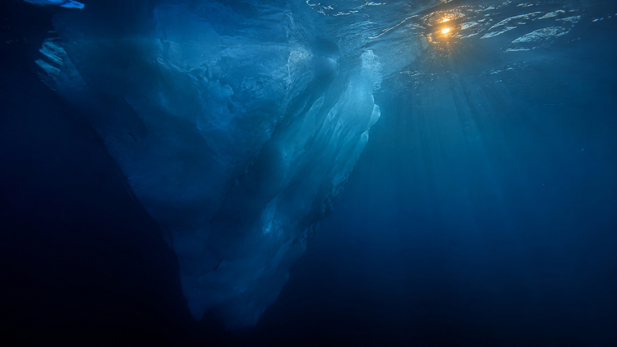 Vista submarina de la enorme masa sumergida de un iceberg, con haces de luz azul y un destello cálido del sol en la superficie.